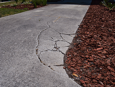 A concrete driveway with large cracks and visible damage along one edge, next to a garden bed covered with red wood mulch, may benefit from professional concrete driveway repair by a trusted residential concrete company.
