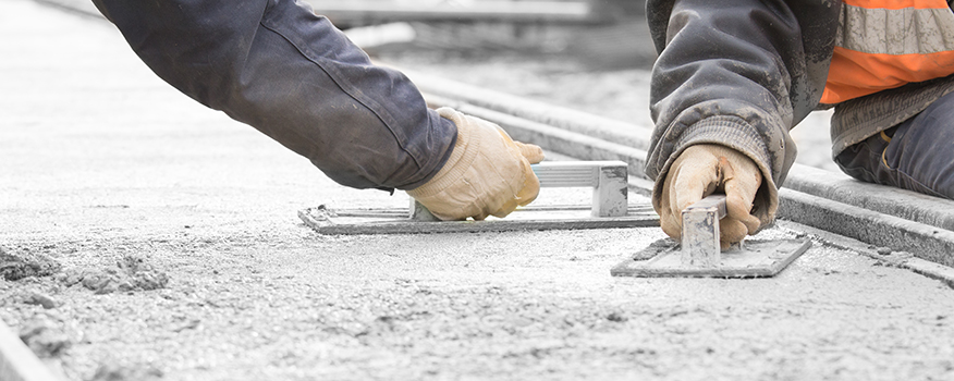 Two construction workers wearing gloves smooth wet concrete with hand trowels. One, from a residential concrete company, wears a dark jacket; the other, a high-visibility vest. The focus is on their hands and tools shaping the fresh concrete surface.