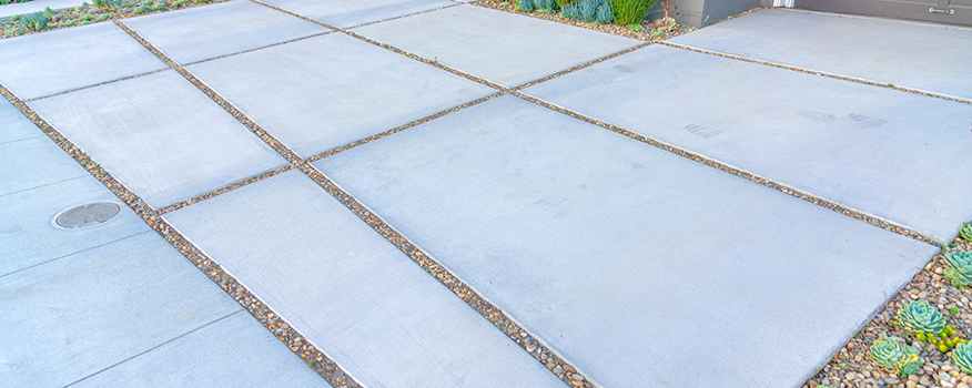 Large concrete slabs arranged in a grid pattern, separated by pebble-filled strips, form a modern driveway bordered by low plants and succulents—a stunning installation by an expert residential concrete company.