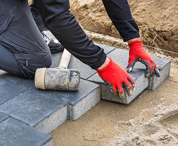 A person wearing red gloves is laying gray paving stones on sand for a walkway or patio, with a mallet nearby, showcasing expert concrete service from a residential concrete company.