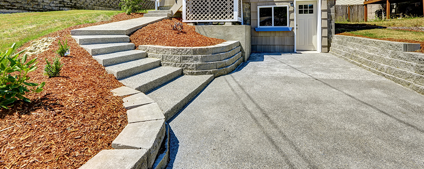 Concrete steps and a curved retaining wall lead up to the entrance of a house with a small porch. The surrounding landscaping features mulch and sparse plants, bordered by concrete and stonework, demonstrating quality concrete service.