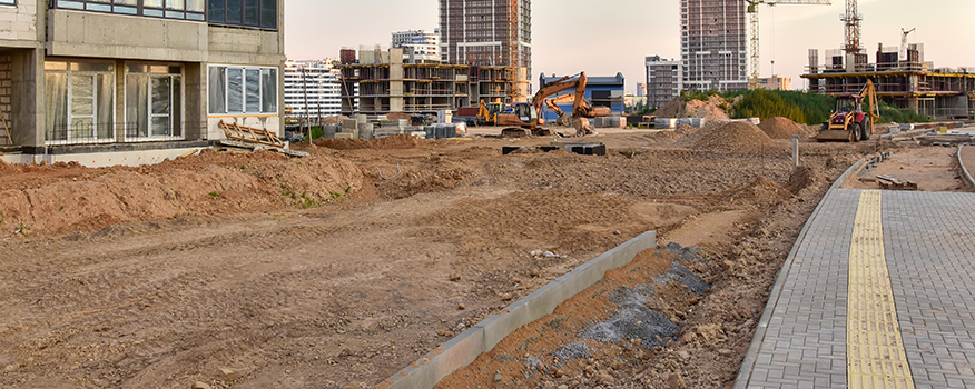 A construction site with dirt, construction vehicles, and unfinished high-rise buildings in the background. A paved sidewalk with a tactile guiding strip, likely crafted by a concrete company Tampa trusts for quality concrete service, runs on the right side of the image.