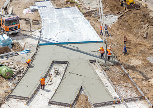Aerial view of construction workers from a residential concrete company pouring and smoothing concrete into a large form shaped like the letter M at a busy construction site.