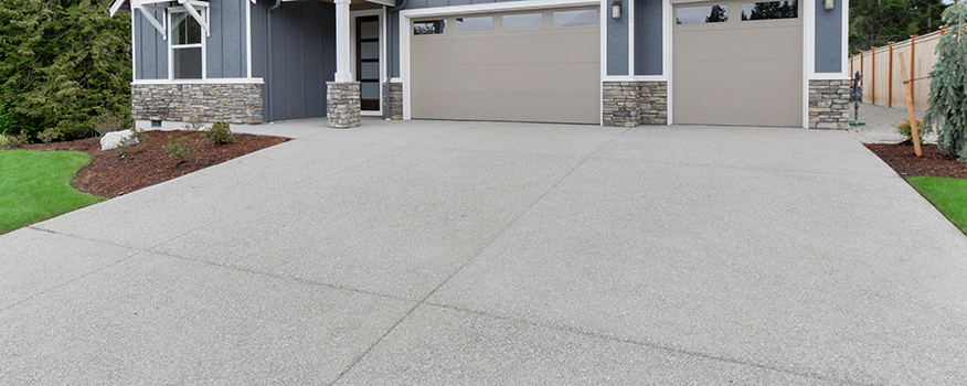 Wide view of a clean, light gray concrete driveway by a leading residential concrete company leading to a modern house with two garage doors, stone accents, green lawn, and landscaped garden beds.