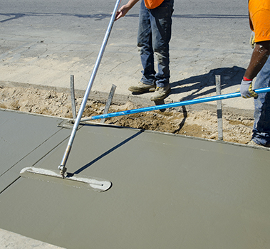 Two workers in orange shirts smooth freshly poured concrete on a sidewalk using long-handled tools. This residential concrete company ensures high-quality concrete service as the wet material is carefully finished near the edge of the street.