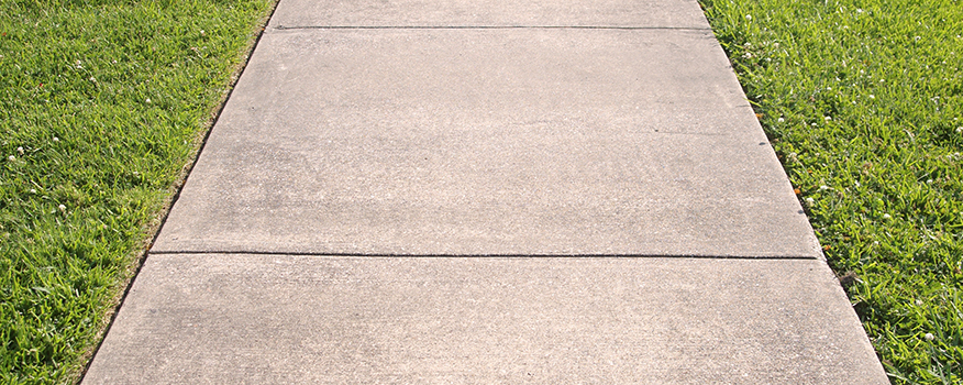 A straight concrete sidewalk with visible seams runs through neatly trimmed green grass on both sides, showcasing the quality you’d expect from a top concrete company Tampa trusts, all under a bright, sunny sky.