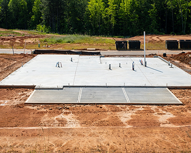 A freshly poured concrete foundation slab with plumbing pipes and conduit emerging, set on a cleared, reddish dirt construction site surrounded by green trees—crafted by a trusted concrete company Tampa residents rely on.