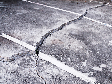 A large crack runs through the surface of a paved road or parking lot, crossing over white painted lines and causing visible damage that may need expert concrete service.