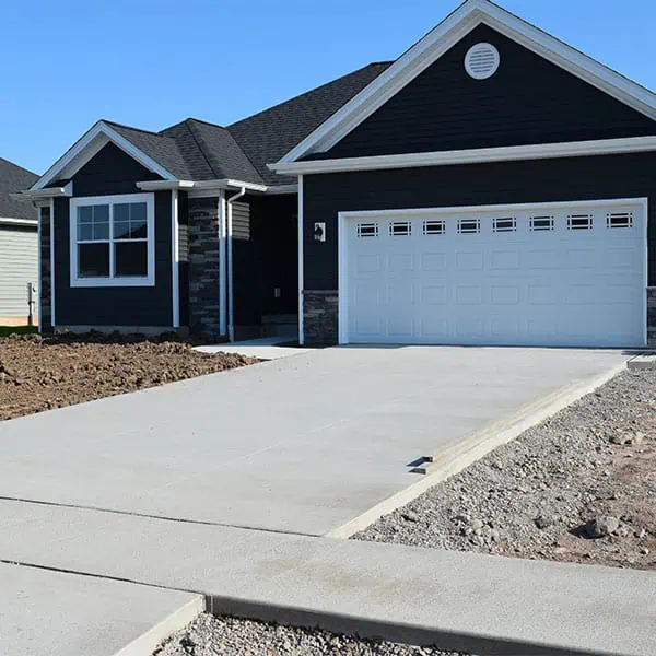 residential-concrete A newly constructed black house with white trim and a double garage door sits behind a fresh concrete driveway, expertly crafted by a leading residential concrete company. The yard is unfinished, with bare soil and gravel surrounding the driveway.