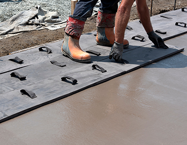 Two workers wearing boots and gloves stand on large rubber mats with handles, placed on freshly poured concrete at a construction site. As part of a residential concrete company, they ensure the surface stays smooth during the concrete pumping process.