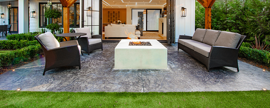 Modern outdoor patio with a central fire pit, cushioned wicker seating, and a sleek concrete floor—expertly installed by a trusted concrete company Tampa—that flows seamlessly into green grass and a stylish indoor living space.