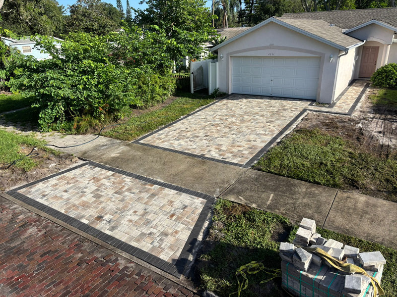 Newly installed driveway and walkway made of rectangular stone pavers with a dark border next to a white house with a two-car garage, expertly completed by a residential concrete company. Stacked pavers and construction materials are visible in the corner.