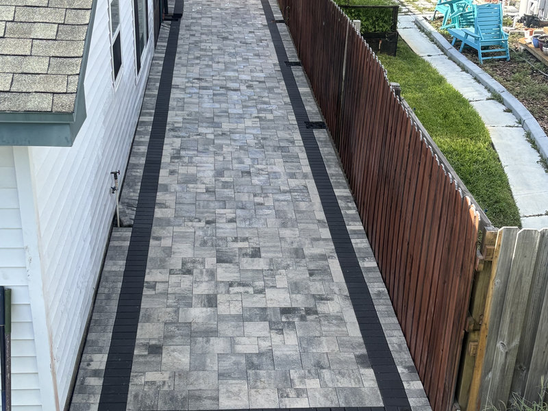 A paved walkway with light gray and dark gray rectangular stones runs alongside a white house and wooden fence—crafted by a leading residential concrete company, with a yard and blue chairs visible in the background.