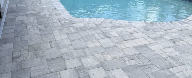 Rectangular swimming pool with clear blue water, bordered by a large patio crafted by a residential concrete company. Light gray rectangular and square stone pavers create a geometric pattern, with a screen enclosure and greenery in the background.