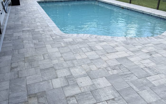 Rectangular swimming pool with clear blue water, bordered by a large patio crafted by a residential concrete company. Light gray rectangular and square stone pavers create a geometric pattern, with a screen enclosure and greenery in the background.
