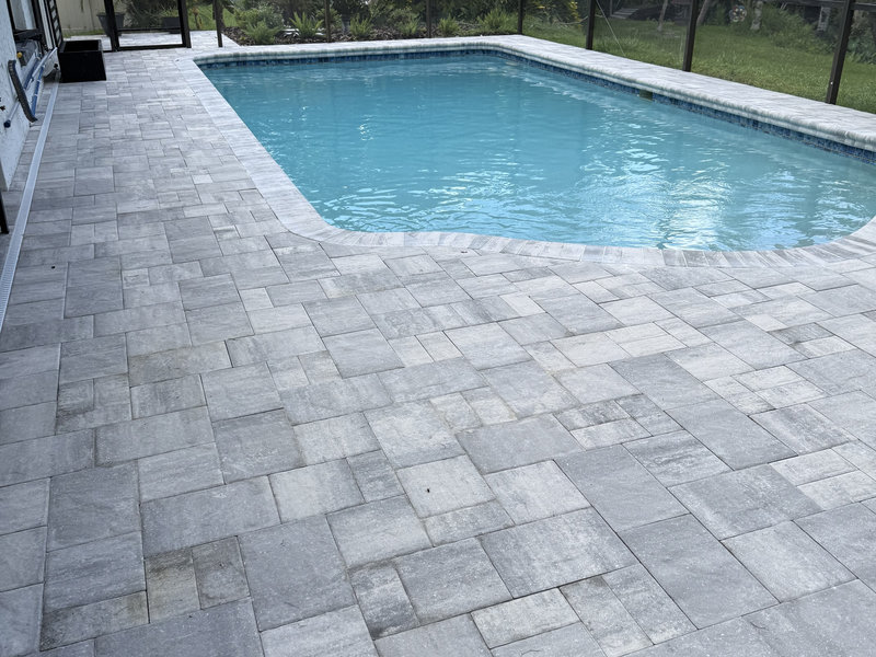IMG_3922 Rectangular swimming pool with clear blue water, bordered by a large patio crafted by a residential concrete company. Light gray rectangular and square stone pavers create a geometric pattern, with a screen enclosure and greenery in the background.