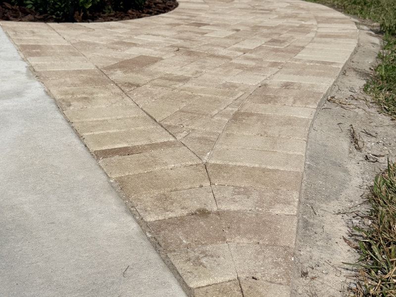 A curved walkway made of rectangular tan pavers, arranged in a herringbone pattern, meets a concrete path—expertly installed by a residential concrete company—on one side and grass on the other.