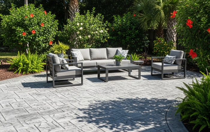 A modern outdoor patio with gray cushioned seating, a coffee table, and armchairs sits on a stamped concrete surface crafted by a top concrete company Tampa, surrounded by lush greenery, palm trees, ferns, and red hibiscus flowers.